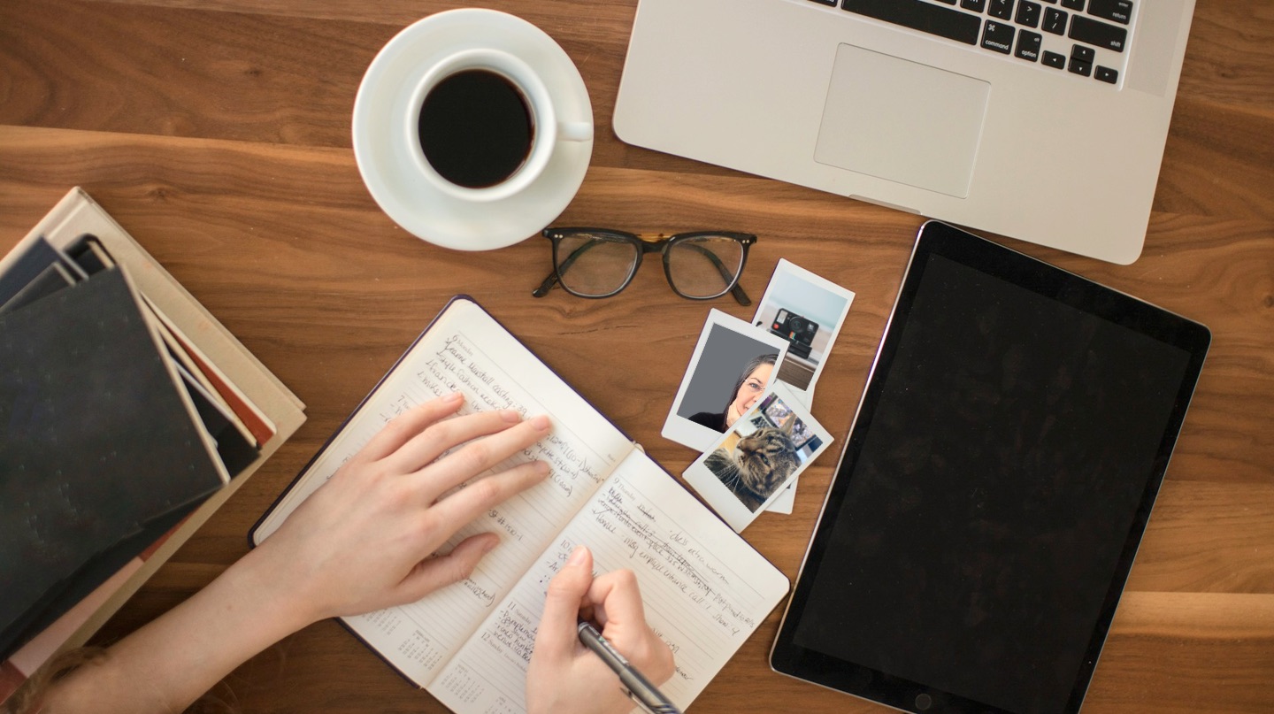 Overhead view of a dark wood desk with a laptop, white cup and saucer with coffee in it, glasses, photos, notebooks, and a pair of hands writing in an open notebook.