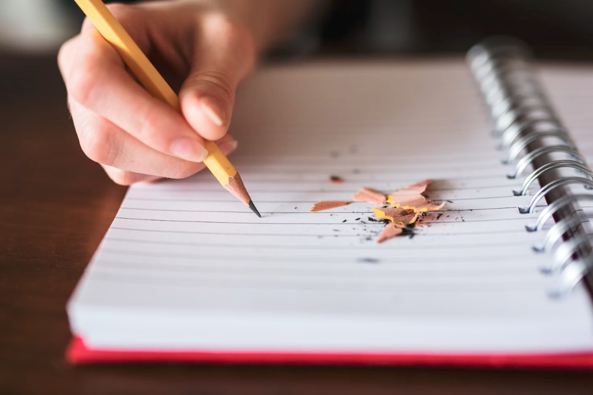 Hand with a yellow pencil writing in a lined spiral notebook. 
