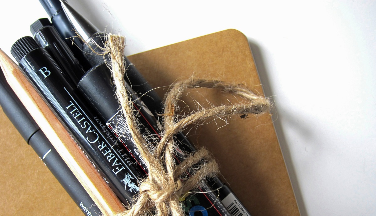 A bundle of pens and markers tied in a string sit on a manila notebook.